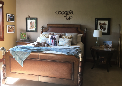 Scrimshaws Portfolio of cowgirl bedroom design with a brown bed frame including pictures of old boots, saddle, and “Cowgirl Up” horseshoe artwork.