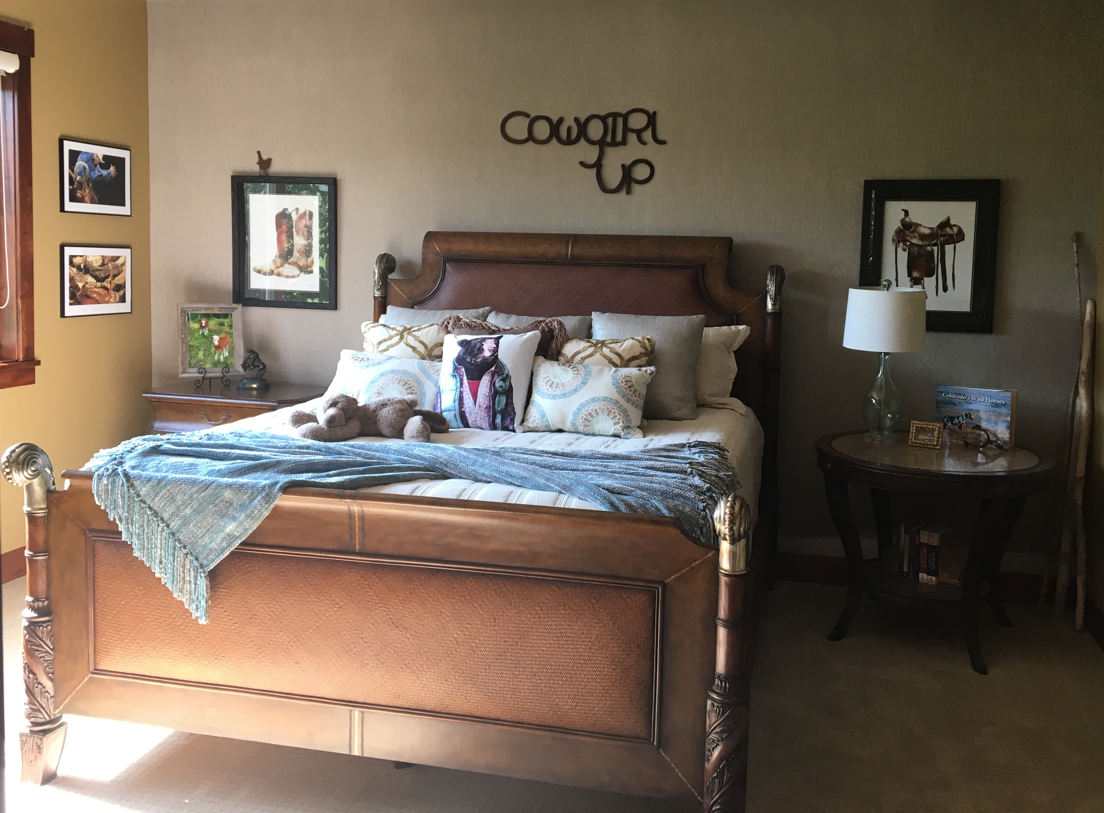 Scrimshaws Portfolio of cowgirl bedroom design with a brown bed frame including pictures of old boots, saddle, and “Cowgirl Up” horseshoe artwork.